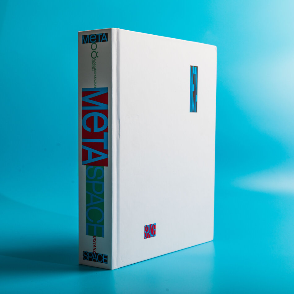 A big white book with a white cover and the title ’Meta space’. The word ’meta’ is inscribed in a red rectangle, while the word ’space’ is inscribed in a smaller green rectangle. The book appears to be floating in a blue background with no visible surroundings.