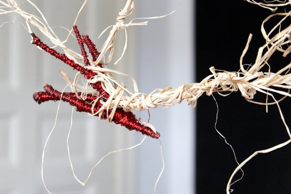 A close-up detail of an artwork featuring raffia, branches, and red metallic enameled wire. The composition shows intertwined strands of raffia and branches, with the red wire adding a striking contrast.